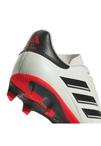 Botín de fútbol blanco con franjas negras y acentos rojos. Presenta un acabado suave y texturizado con un talón acolchado para mayor soporte.