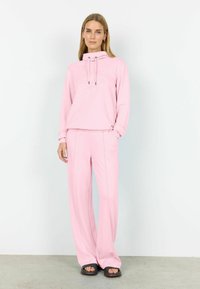 Svetloružová súprava pozostávajúca z mikiny s kapucňou a šnúrkami a voľného strihu, kombinovaná so širokými nohavicami s vreckami.