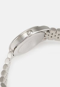 Montre en acier inoxydable avec un boîtier rond et un bracelet texturé. Dispose d'une couronne avec un branding sur le côté. La couleur est principalement argentée.