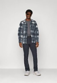 Flis jakna v temno sivi barvi s karo vzorcem, kombinirana s sivo srajco in temnimi hlačami. Model nosi sončna očala in sive čevlje.