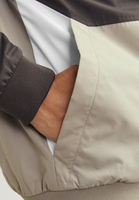 Lightweight, color-blocked jacket featuring a gray top section, beige body, elastic cuffs, and a side pocket with a white interior lining.