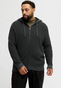 Haut zippé gris foncé avec un motif tricoté texturé, capuche avec cordon de serrage et poignets côtelés. Porté sur une chemise vert olive.