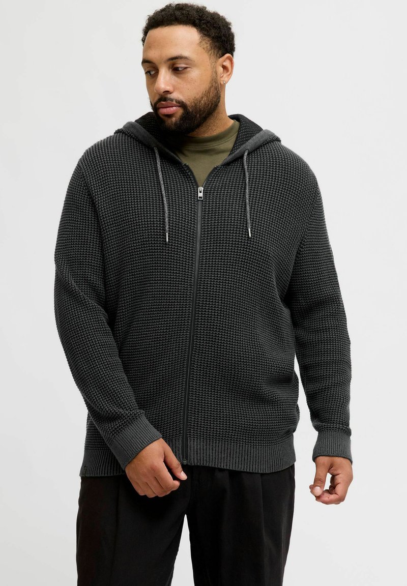 Haut zippé gris foncé avec un motif tricoté texturé, capuche avec cordon de serrage et poignets côtelés. Porté sur une chemise vert olive.