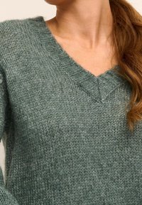 Pull en tricot vert avec un col en V, présentant un motif texturé et des bords en côtes, fabriqué à partir d'un matériau doux et moelleux.