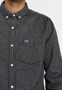 Camisa de franela gris oscuro con cuello abotonado, botones blancos y un bolsillo en el pecho izquierdo con una etiqueta de marca azul marino.