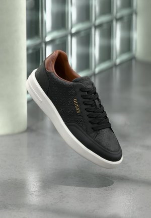 Sneaker in pelle nera con motivo a logo impressa, collo marrone, suola bianca e scritta dorata "GUESS", su uno sfondo di piastrelle moderne.