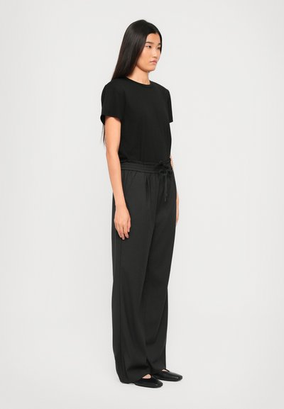 Young woman in black short-sleeve shirt, wide black drawstring pants, and black flats, standing sideways against a plain white background.