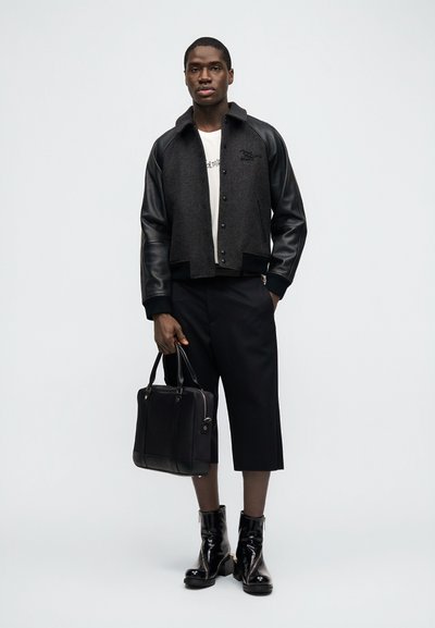 Άνδρας που φοράει μαύρο και γκρι varsity μπουφάν, λευκό T-shirt, μαύρο παντελόνι μέχρι τον αστράγαλο, μαύρα μποτάκια αστραγάλου, κρατώντας μια μαύρη δερμάτινη τσάντα.