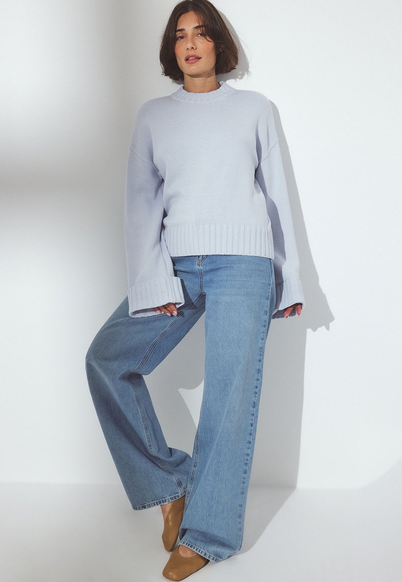 Kvinna som bär en ljusblå oversized tröja och vida blå jeans, stående med ett ben böjt mot en vit vägg.