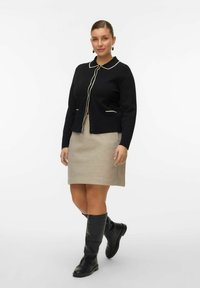Sort cardigan med hvid kant, knaplukning fortil, parret med en beige nederdel og sort støvler til knæet. Enkel, skræddersyet silhuet.