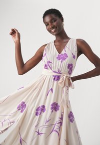 Abito floreale con sfondo beige e vivaci fiori viola, scollatura a V profonda e cintura con lacci in vita per una silhouette fluida.