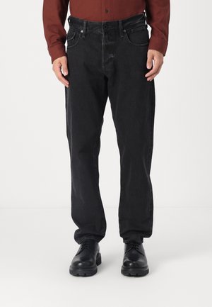 Mann trägt schwarze Jeans mit geradem Bein, schwarze Lederschuhe und ein dunkelrotes Hemd mit langen Ärmeln und Knöpfen, steht vor einem weißen Hintergrund.