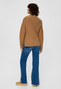 Brauner Strickpullover mit entspanntem Schnitt, weiten Ärmeln und einem leicht längeren Rücken. Kombiniert mit blauen, ausgestellten Jeans und hellbraunen Schuhen.
