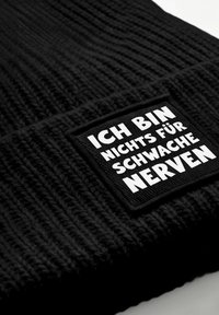 Schwarze, gestrickte Mütze mit strukturiertem Muster und einem rechteckigen, bestickten Aufnäher mit weißem deutschen Text.