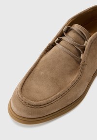 Chaussure marron en daim avec une texture lisse, des détails cousus et des lacets plats, présentant un bout arrondi et une semelle en caoutchouc épaisse couleur tan.