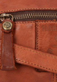 Brown leather bag detail featuring a bronze zipper, anchor button accent, and visible stitching on a smooth textured surface.