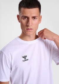 Balta kokvilnas t-kreklu ar apaļu kaklu. Iezīmēts ar mazu melnu izšūtu logotipu uz krūtīm. Gluda tekstūra, standarta piegriezums.