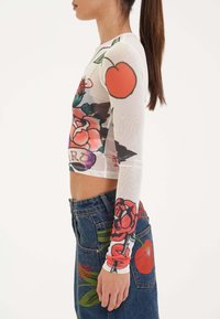 Transparante witte blouse met lange mouwen, versierd met kleurrijke bloemen- en fruitprints, gecombineerd met een blauwe denim rok die soortgelijke ontwerpen heeft.
