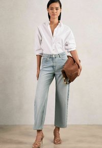 Gaiši zili augstās jostas bikses ar brīvu piegriezumu, apvienotas ar baltu pogājamu krekli un brūnām ādas somām ar siksniņām. Tur rokās brūnu ādas somu.