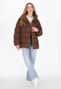 Bruine pufferjack met doorgestikt ontwerp, hoge kraag en drukknoopsluiting, gecombineerd met lichtblauwe jeans en witte sneakers.