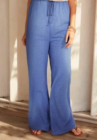 Blauwe brede broek van gestructureerde stof met een drawstring taille en een subtiele glans. Gecombineerd met een paar bruine slippers op een houten vloer.