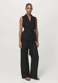 Donna in piedi che indossa un blazer senza maniche nero con cintura e pantaloni neri a gamba larga, con sandali a piedi nudi su uno sfondo leggero e semplice.