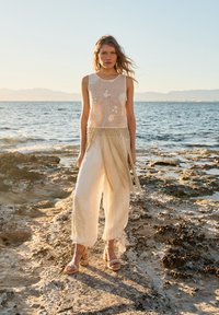 Woman standing on rocky shore wearing a sleeveless mesh top, flowing wide-leg pants, and woven heeled sandals by the sea at sunset.