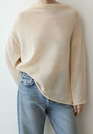 Personne portant un pull en maille beige ample rentré dans un jean taille haute bleu clair, debout devant un fond clair uni.