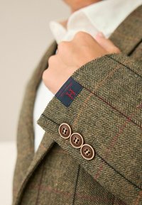 Houndstooth tweedjacka med tre bruna knappar och marinblå etikett. Har ett strukturerat tyg med jordnära toner och en subtil rutmönstring.