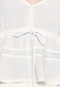 Blusa bianca in tessuto leggero e testurizzato, con scollo a V, vita arricciata e motivo decorativo in pizzo con cordoncino.