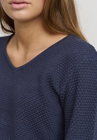 Navyblauer Strickpullover mit einem strukturierten, waffleartigen Muster, mit V-Ausschnitt und langen Ärmeln, der eine lockere Passform betont.