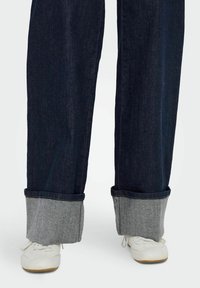 Tumšas džinsa bikses ar sarullētām piedurknēm, kas izcelta ar kontrastējošu gaiši pelēku audumu, apvienotas ar baltām sporta kedām.