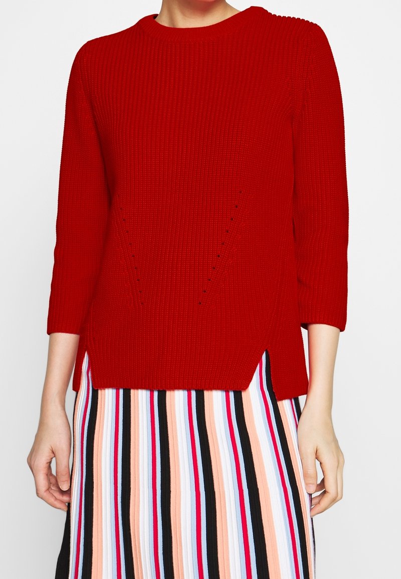 Roter gerippter Pullover mit Dreiviertelärmeln; Seitenschlitze am Saum; kombiniert mit einem mehrfarbigen gestreiften Rock mit vertikalen Linien.