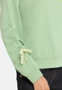 Jasnozielony sweter z gromadzonymi rękawami, ozdobiony żółtymi wiązaniami przy nadgarstkach. Miękka tekstura, ściągacze przy mankietach i przytulny design.