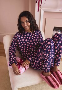 Jūras spēku pidžamu komplekts ar rozā zvaigžņu rakstu, garām piedurknēm un biksēm. Mīkstas rozā pūkainas mājas čības. Dāvanu kaste rokās ar rozā lentu. Krēmkrāsas fons.