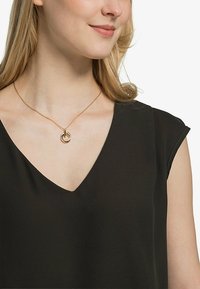 Guldfarvet halskæde med sammenflettede cirkulære vedhæng på en delikat kæde, båret med en sort v-hals top, som fremhæver dets enkle design.