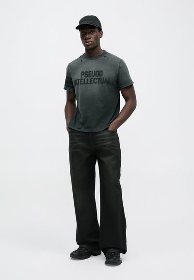 Schwarzes Grafik-T-Shirt mit dem Aufdruck "PSEUDO INTELLECTUAL", ausgefranste Kanten, kombiniert mit lockeren schwarzen Jeans und schwarzen Turnschuhen.