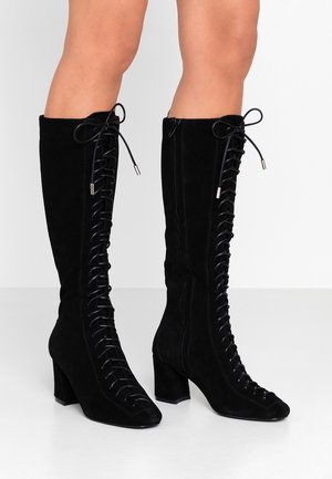 Black suede knee-high boots with block heels, front lace-up design, and side zippers worn on bare legs against a plain background.