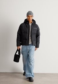 Pufferjakke i svart med hette og logo, kombinert med lyseblå jeans og svarte støvler. Tilbehør inkluderer en grå lue og en liten veske.