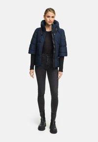 Vrouw in een marineblauwe pufferjack met korte mouwen over een zwarte long-sleeve top, zwarte skinny jeans en zwarte enkellaarsjes, staande op een witte achtergrond.