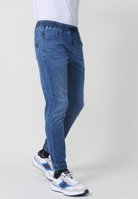 Pantalones jogger de denim en azul medio, con cintura ajustable mediante cordón, puños elásticos y bolsillos laterales con detalles de costura visibles.