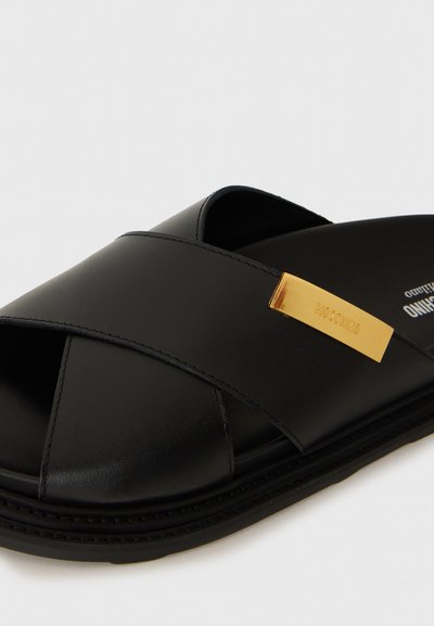 Svarta lädersandaler med korsade remmar och en rektangulär gul metallemblem graverad med "MOSCHINO."