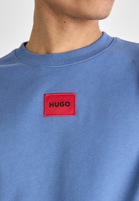Felpa azzurra con collo a giro, dotata di una toppa rettangolare rossa con la scritta "HUGO" in nero. Texture liscia, design casual.