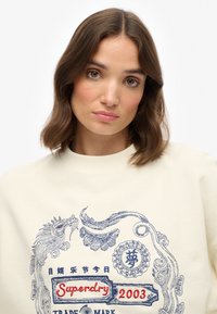 Witte sweatshirt met blauwe borduursels van een draak en de tekst "Superdry 2003" met ingewikkelde decoratieve elementen en een fijne textuur.