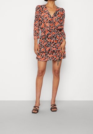 Woman wearing a black dress with orange and white floral patterns, three-quarter sleeves, and black strappy sandals, standing against a plain wall.