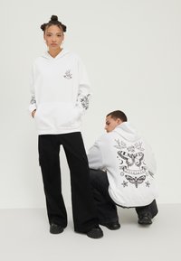 Hvit hettegenser med frontlomme, med svarte grafiske utskrifter av fugler og blomsterdesign på ermene. Svarte vide bukser og kraftige sko.
