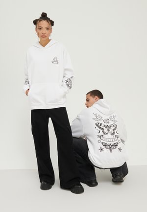 Sudadera blanca con bolsillos frontales, que presenta estampados gráficos en negro de aves y diseños florales en las mangas. Pantalones anchos negros y zapatos gruesos.