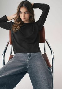 Maglietta nera a maniche lunghe con scollatura rotonda, abbinata a jeans grigi chiari a vita alta. Modella seduta su una sedia in cromo con dettagli in pelle marrone.