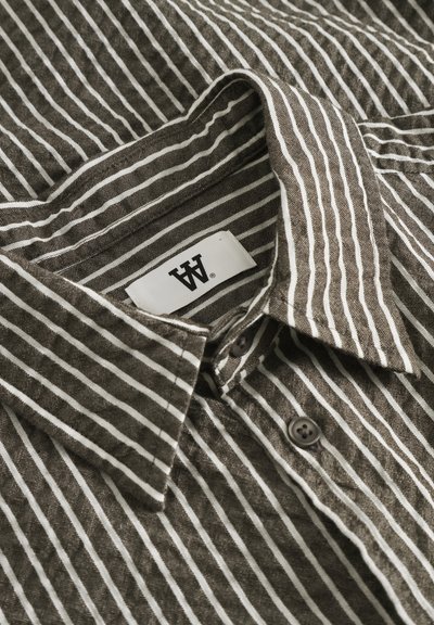 Chemise boutonnée rayée marron et blanche avec col, présentant une texture tissée et deux boutons marron près du cou. Étiquette de marque visible.