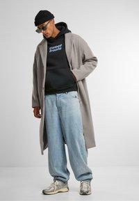 Gray long coat over a black hoodie featuring blue text, paired with light blue wide-leg jeans and gray sneakers. Accessories include a black beanie and green sunglasses.